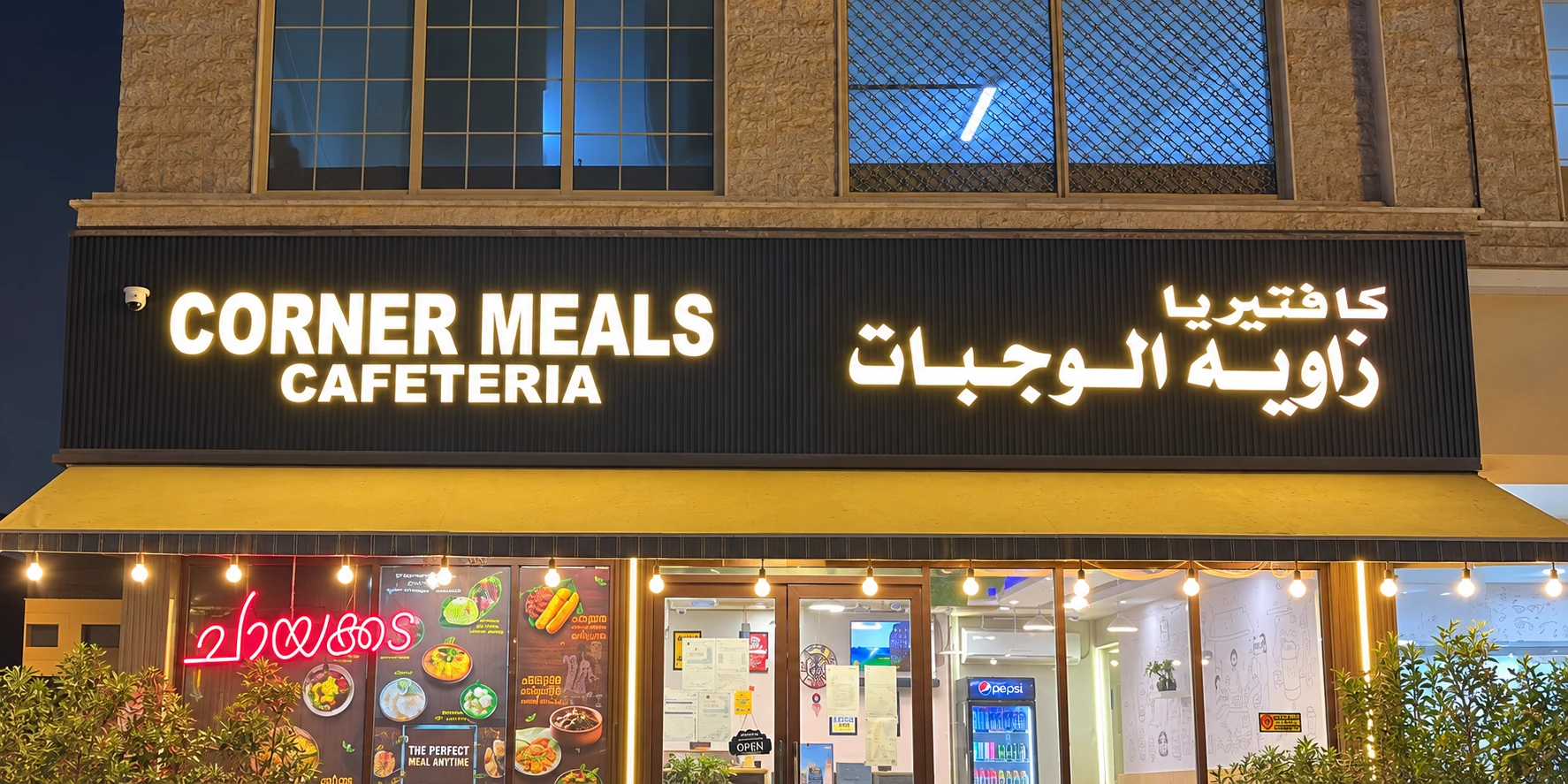 Corner Meals Cafeteria's illuminated English and Arabic storefront sign above its mustard awning.